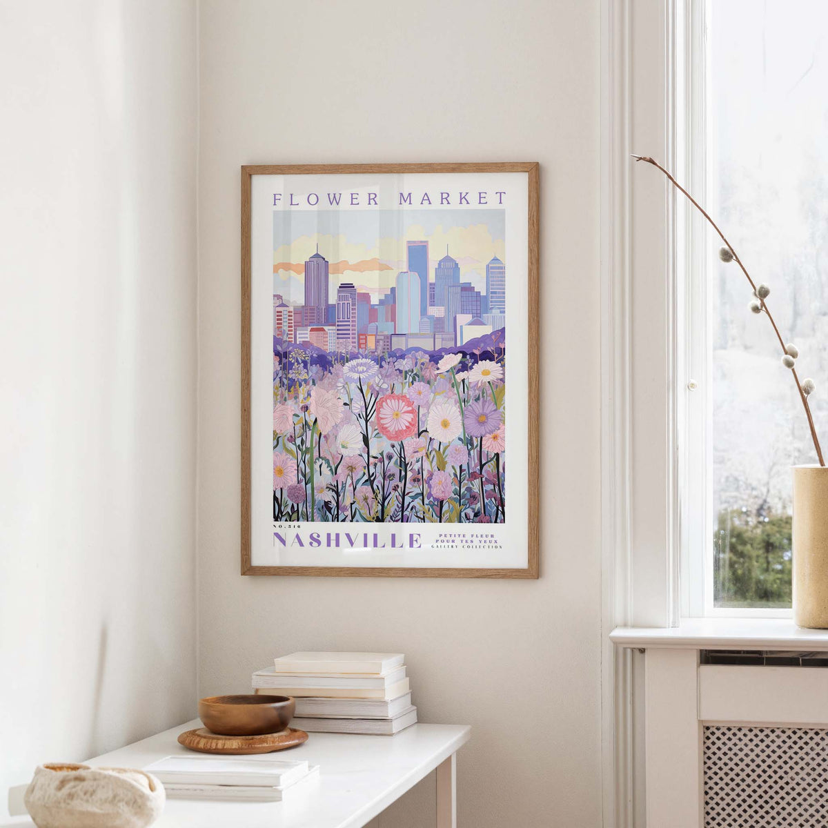 a framed poster of a cityscape with a field of flowers in the foreground, hanging on a wall in a room with a window and a table with books and a vase.