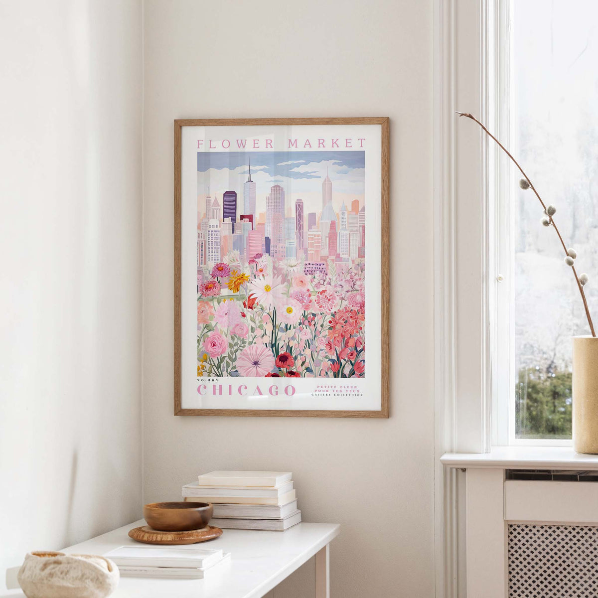 a framed poster of a cityscape with a flower market in Chicago, hanging on a wall in a room with a window and a small table with books and a bowl.