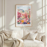a framed poster of a colorful garden scene hanging on a wall above a white couch with a blanket draped over it.