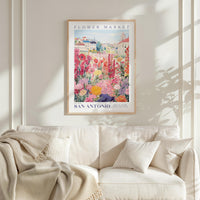 a framed poster of a colorful garden scene hanging on a wall above a white couch with a blanket draped over it.
