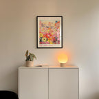a white cabinet with a potted plant and a lamp on top, set against a white wall. Above the cabinet, there is a framed poster of a colorful floral scene.