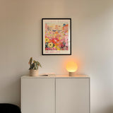 a white cabinet with a potted plant and a lamp on top, set against a white wall. Above the cabinet, there is a framed poster of a colorful floral scene.
