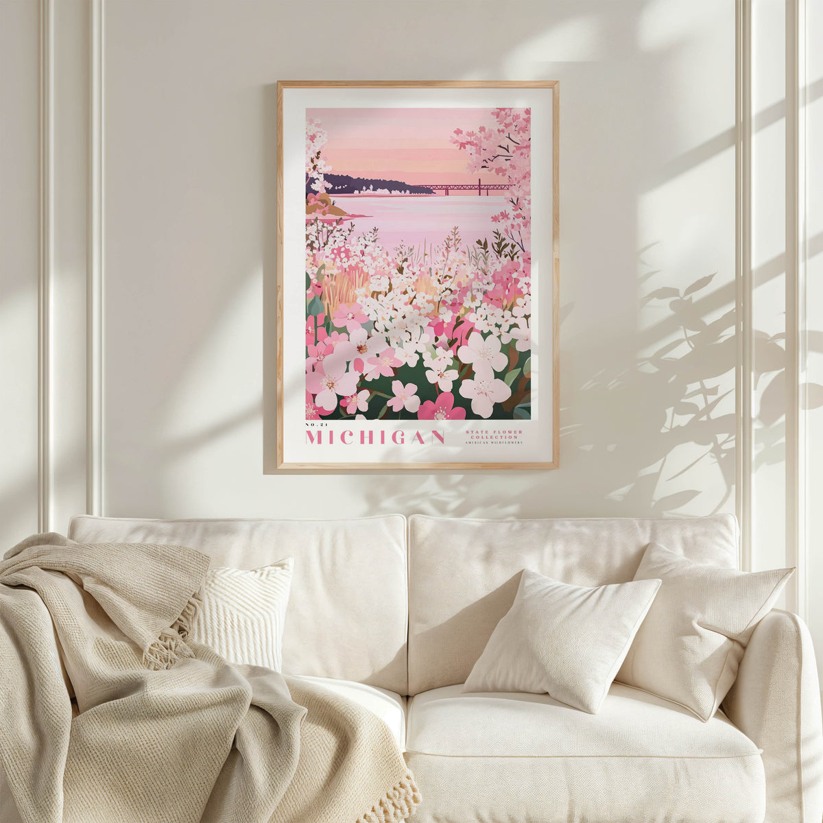 a framed poster of a serene landscape with pink flowers and a sunset, hanging on a white wall above a white couch with a blanket draped over it.