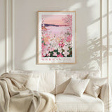 a framed poster of a serene landscape with pink flowers and a sunset, hanging on a white wall above a white couch with a blanket draped over it.