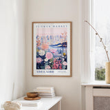 a framed poster of a cityscape with a floral design, hanging on a wall in a room with a window and a small table with books and a bowl on it.