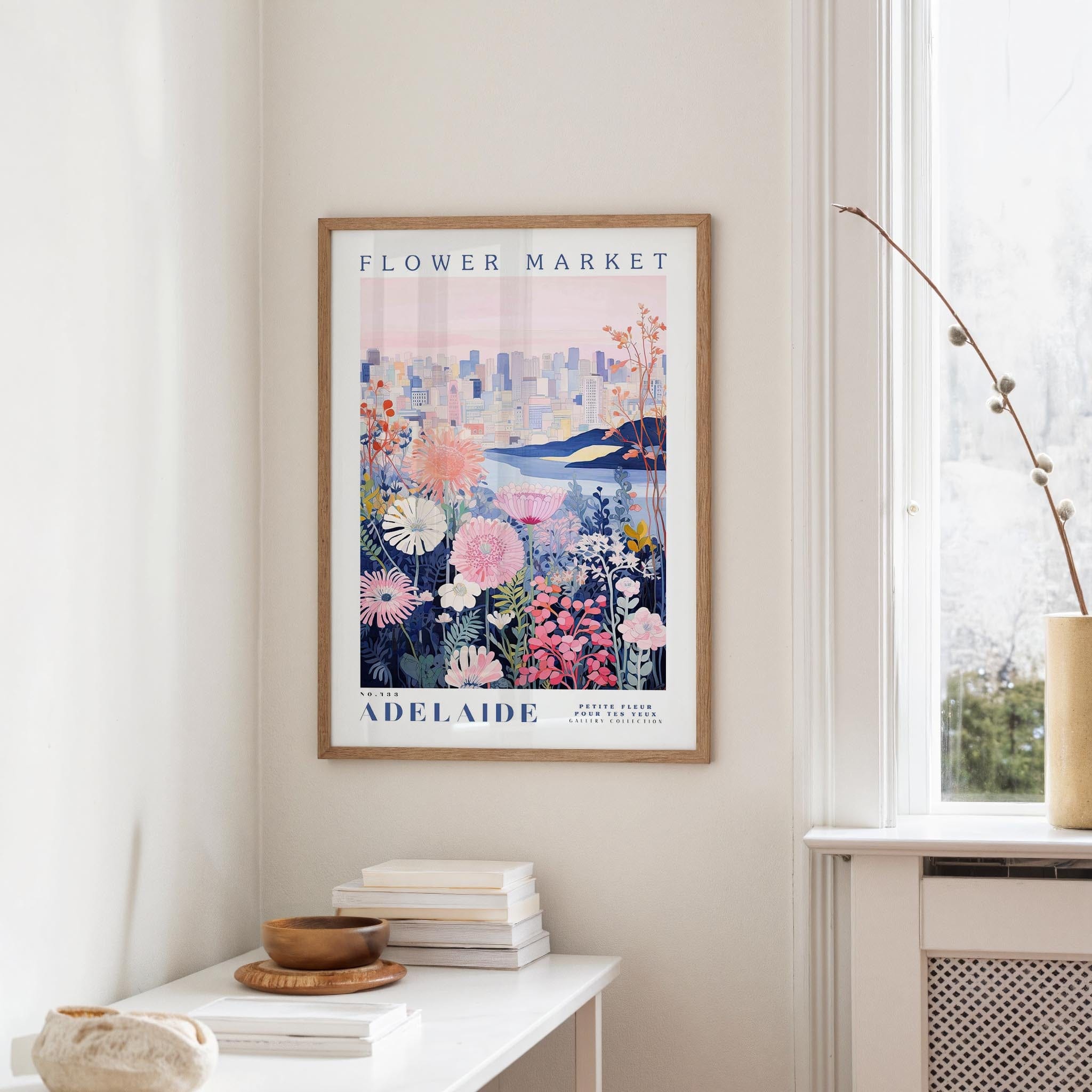 a framed poster of a cityscape with a floral design, hanging on a wall in a room with a window and a small table with books and a bowl on it.