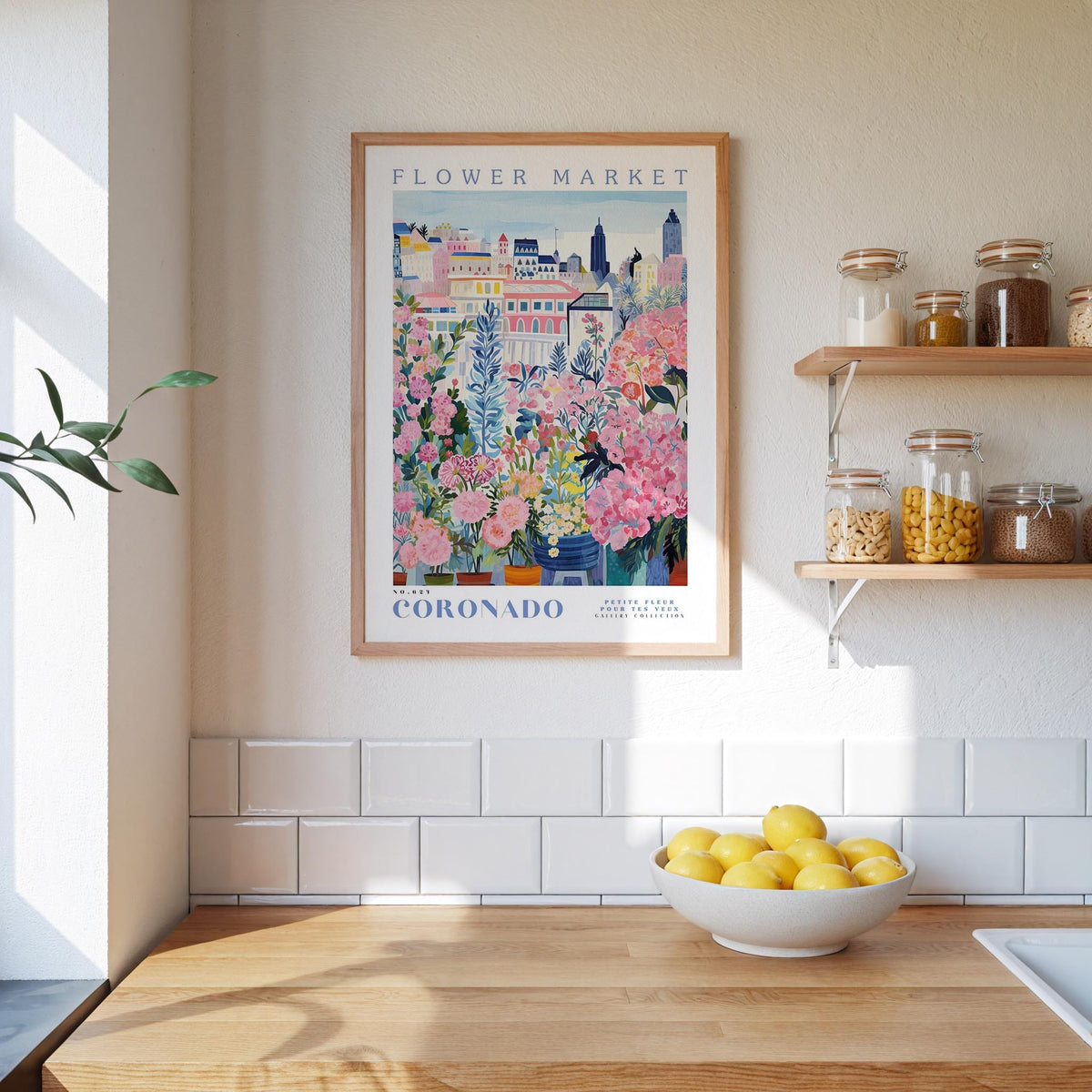 a kitchen scene with a framed poster of a colorful floral market in Coronado, California, hanging on the wall above a wooden counter. The poster is surrounded by various kitchen items, such as jars, a bowl of lemons, and a plant.