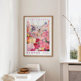 a framed poster of a colorful floral scene hanging on a wall, with a white table and a vase of flowers in the foreground.