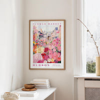 a framed poster of a colorful floral scene hanging on a wall, with a white table and a vase of flowers in the foreground.