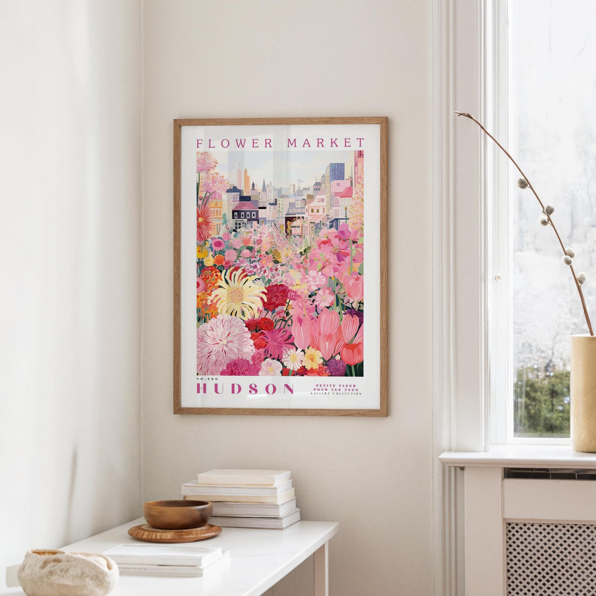 a framed poster of a colorful floral scene hanging on a wall, with a white table and a vase of flowers in the foreground.