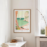 a framed poster of a beach scene with a boat, palm trees, and a rocky cliff, hanging on a wall in a room with a window and a small table with books and a bowl.
