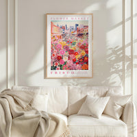 a framed poster of a vibrant flower market in Frisco, California, hanging on a white wall above a white couch with a blanket draped over it.