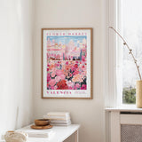 a framed poster of a colorful floral scene hanging on a wall, with a white table and a vase of flowers in the foreground.