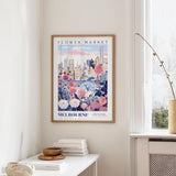 a framed poster of Melbourne's flower market, with a cityscape and colorful flowers on the poster. The poster is hanging on a wall, and there is a table with books and a vase nearby.