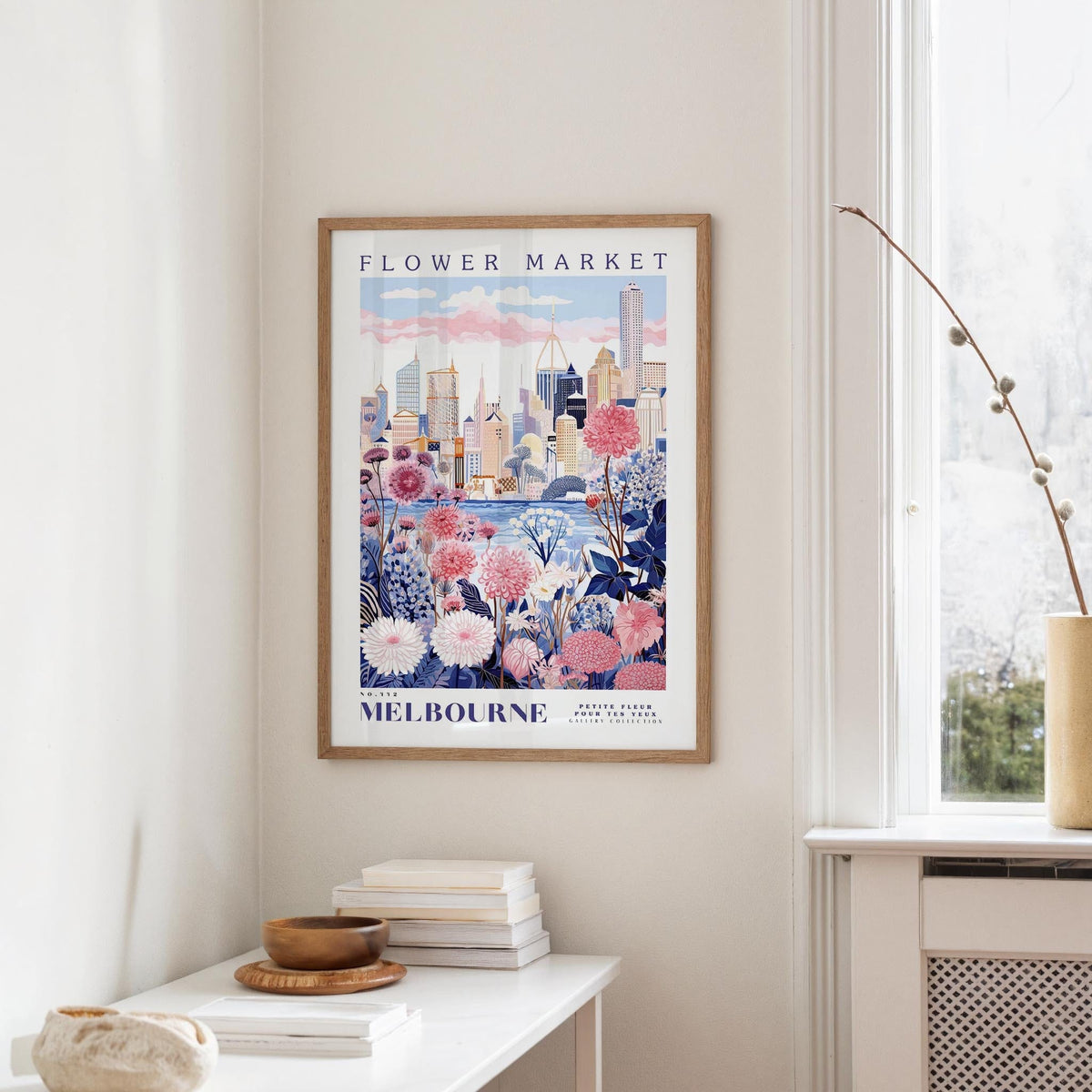a framed poster of Melbourne's flower market, with a cityscape and colorful flowers on the poster. The poster is hanging on a wall, and there is a table with books and a vase nearby.