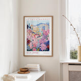 a framed poster of a colorful floral scene hanging on a wall, with a window and a small table with books and a vase in the foreground.