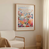 a framed poster of a colorful floral scene hanging on a wall, with a white armchair and a beige vase in the foreground.