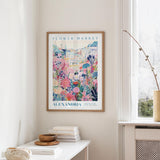 a framed poster of a colorful floral scene hanging on a wall, with a white table and a vase of flowers nearby.
