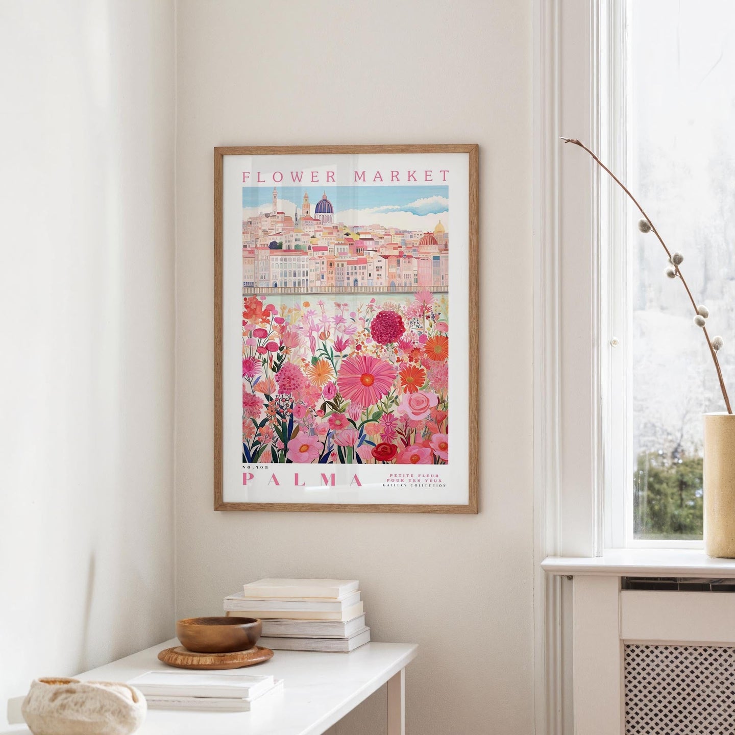 a framed poster of a cityscape with a flower market in the background, hanging on a wall in a room with a window and a small table with books and a bowl on it.