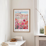 a framed poster of a cityscape with a flower market in the background, hanging on a wall in a room with a window and a small table with books and a bowl on it.