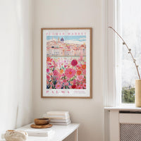 a framed poster of a cityscape with a flower market in the background, hanging on a wall in a room with a window and a small table with books and a bowl on it.