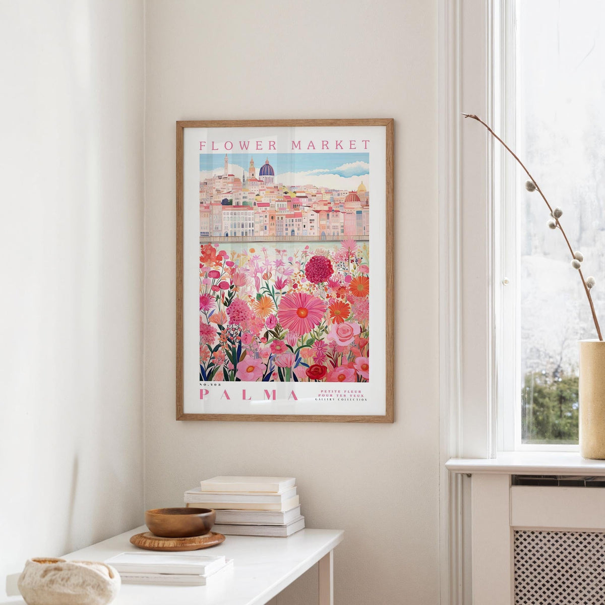 a framed poster of a cityscape with a flower market in the background, hanging on a wall in a room with a window and a small table with books and a bowl on it.