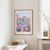a framed poster of the Brisbane Flower Market hanging on a wall, with a window and a small table with books and a vase in the foreground.