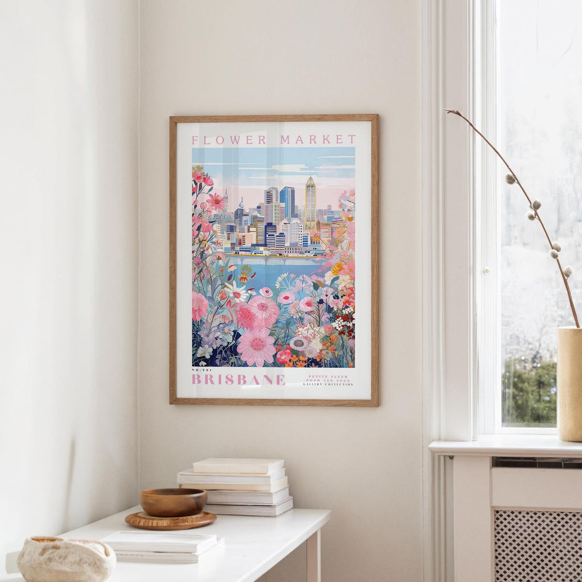 a framed poster of the Brisbane Flower Market hanging on a wall, with a window and a small table with books and a vase in the foreground.