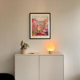 a white cabinet with a potted plant and a lamp on top, and a framed poster of a colorful street scene hanging on the wall above it.