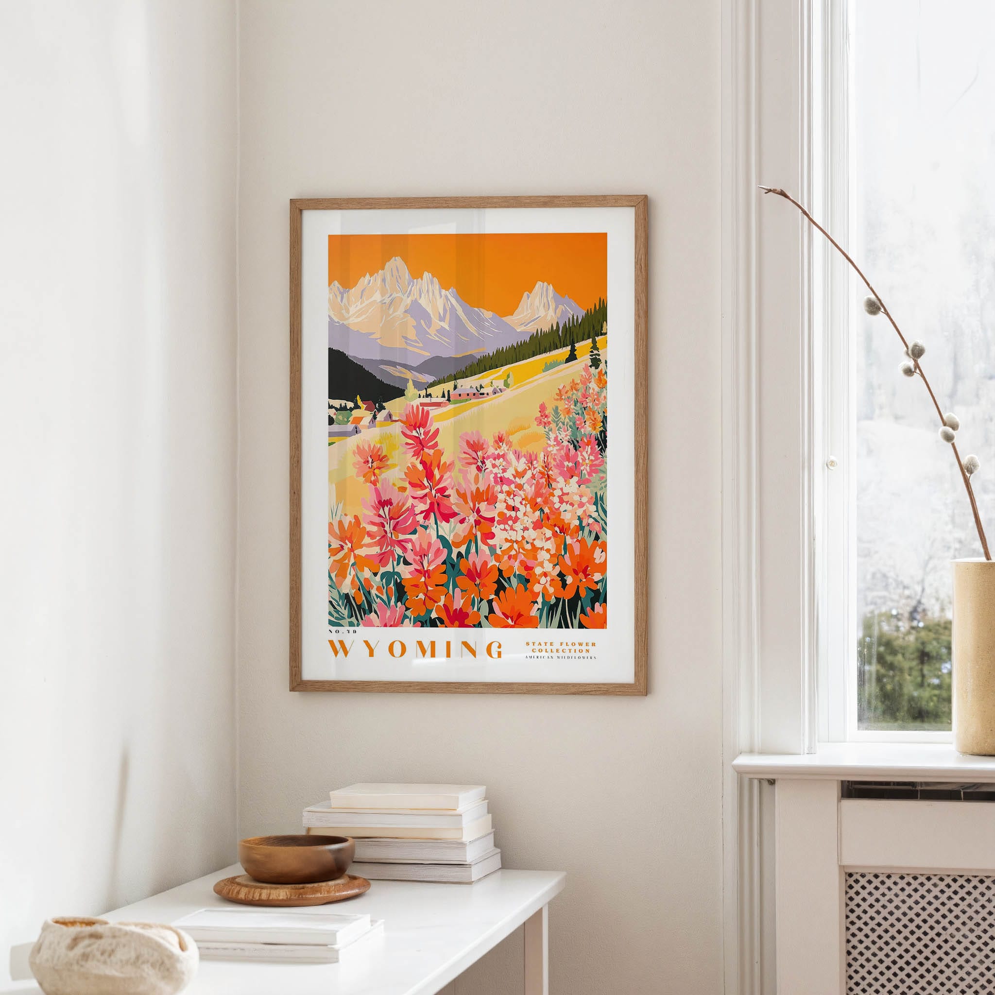 a framed poster of a scenic mountain landscape with a field of colorful flowers, hanging on a wall in a room with a white table and a window.