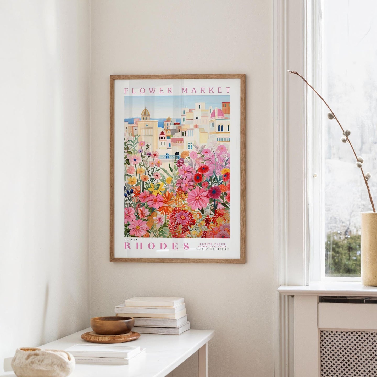 a framed poster of a colorful floral scene hanging on a wall, with a white table and a vase of flowers in the foreground.