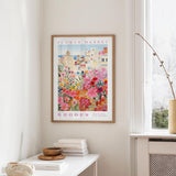 a framed poster of a colorful floral scene hanging on a wall, with a white table and a vase of flowers in the foreground.