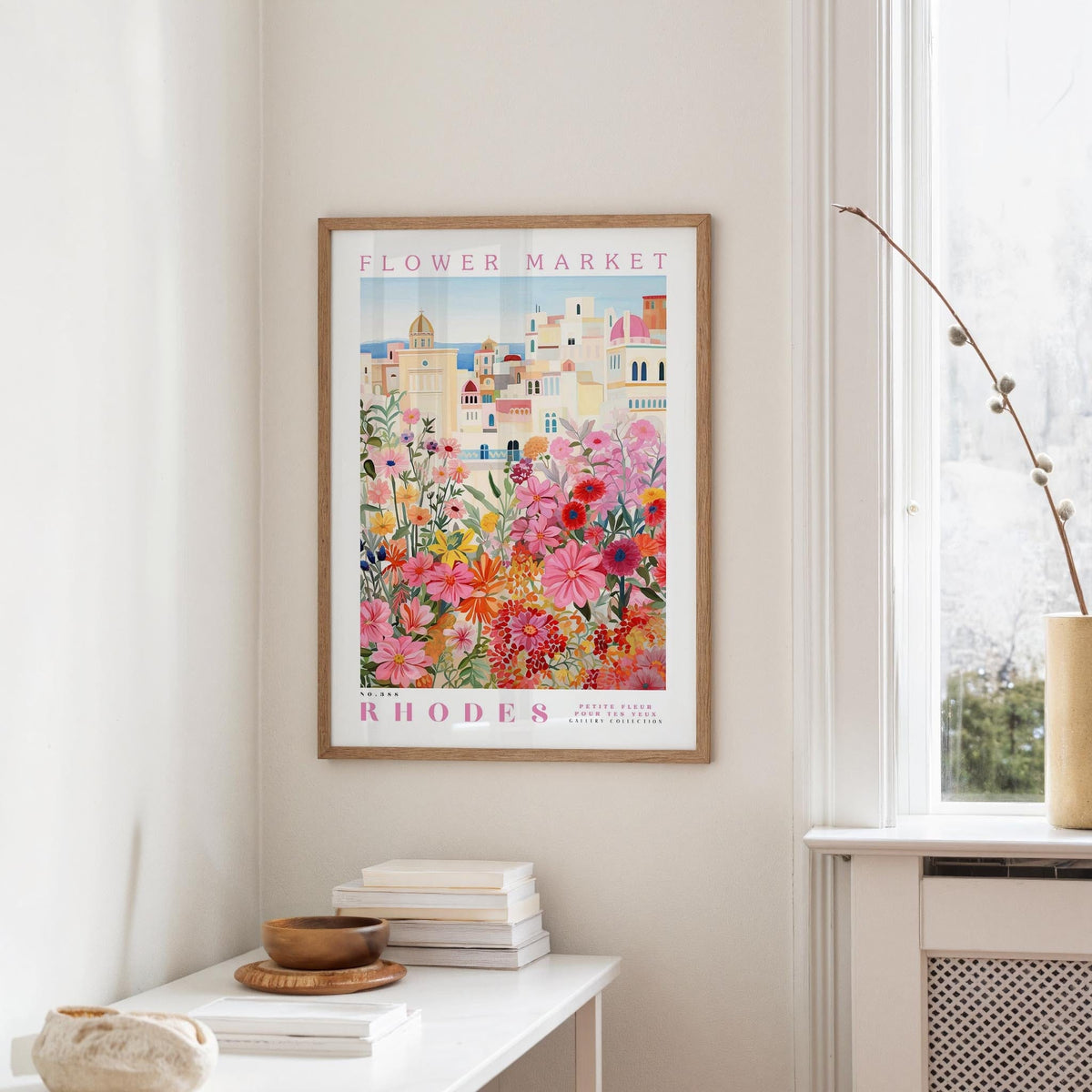 a framed poster of a colorful floral scene hanging on a wall, with a white table and a vase of flowers in the foreground.
