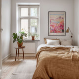 A cozy bedroom with a large bed, a nightstand with a plant, and a framed poster of a cityscape. The room has a warm, inviting atmosphere with a wooden floor and a window overlooking greenery outside.