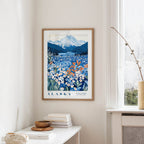 a framed poster of a scenic landscape with mountains, flowers, and a lake, hanging on a wall in a room with a window and a small table with books and a vase.