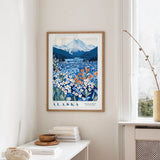 a framed poster of a scenic landscape with mountains, flowers, and a lake, hanging on a wall in a room with a window and a small table with books and a vase.