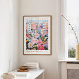a framed poster of a colorful floral scene hanging on a wall, with a white table and a vase of flowers nearby.