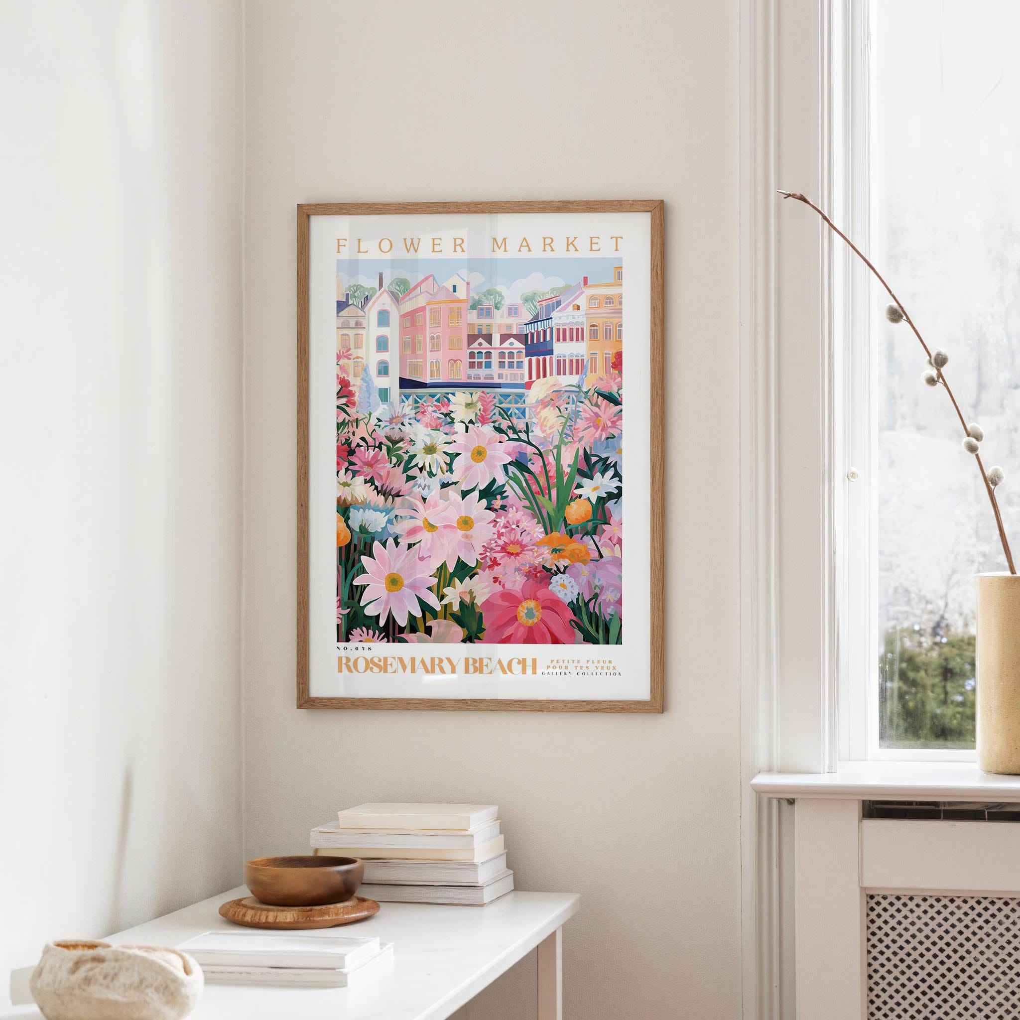 a framed poster of a colorful floral scene hanging on a wall, with a white table and a vase of flowers nearby.