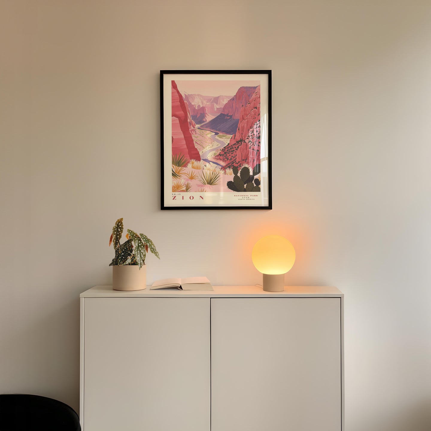 a framed poster of a desert landscape, a potted plant, and a small lamp on a white cabinet.