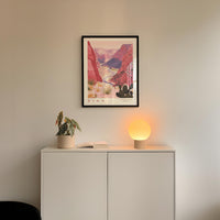 a framed poster of a desert landscape, a potted plant, and a small lamp on a white cabinet.