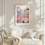 a framed poster of a colorful cityscape with flowers, hanging on a wall above a white couch with pillows.