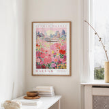 a framed poster of a cityscape with a field of flowers in the foreground, hanging on a wall in a room with a window and a small table with books and a bowl on it.