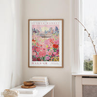 a framed poster of a cityscape with a field of flowers in the foreground, hanging on a wall in a room with a window and a small table with books and a bowl on it.