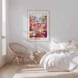 a bedroom with a bed, a woven chair, and a framed poster of a flower market in Greensboro.