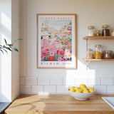 a kitchen scene with a framed poster of a flower market in Bilbao hanging on the wall above a wooden countertop. On the countertop, there is a bowl of lemons and a plant.