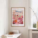 a framed poster of a colorful floral scene hanging on a wall, with a white table and a vase of flowers nearby.