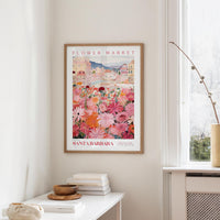 a framed poster of a colorful floral scene hanging on a wall, with a white table and a vase of flowers nearby.