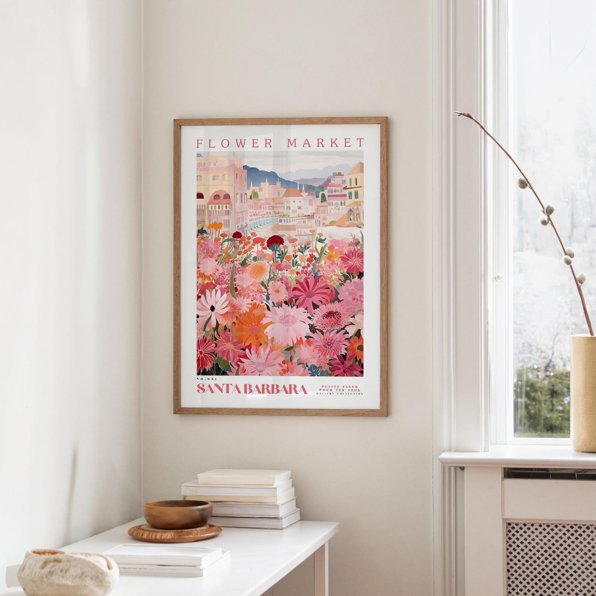 a framed poster of a colorful floral scene hanging on a wall, with a white table and a vase of flowers nearby.