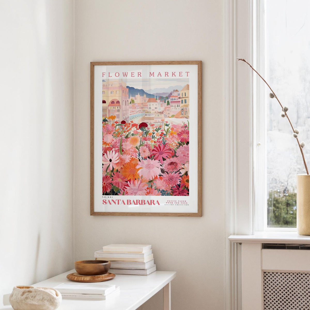 a framed poster of a colorful floral scene hanging on a wall, with a white table and a vase of flowers nearby.