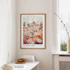 a framed poster of a colorful floral scene hanging on a wall, with a white table and a vase of flowers in the foreground.